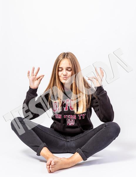 Bubbly blonde young girl in playful oriental pose barefoot with funny expression and closed eyes dressed casually with jeans and sweatshirt