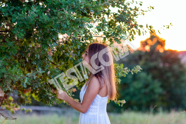 A romantic young girl in profile with long blond hair, dressed in white, at sunset, touches the