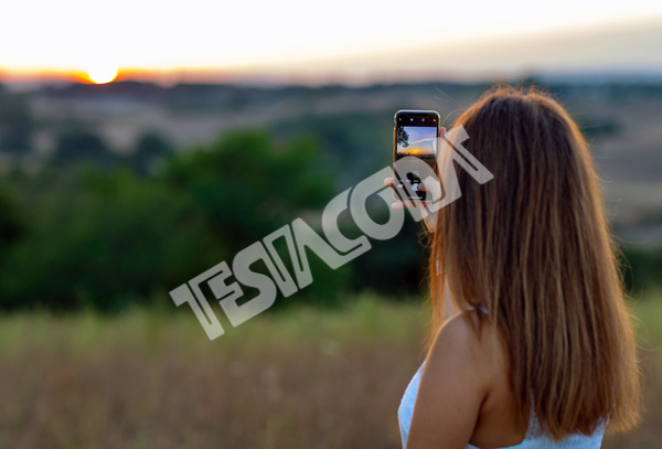 Young girl from behind with long blond hair, dressed in white, takes pictures of setting sun with her mobile phone in countryside